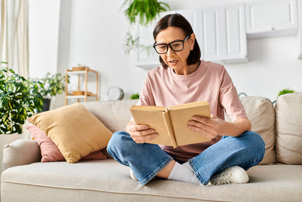Una donna matura in accogliente biancheria da casa seduta su un divano, assorto nella lettura di un libro. - Foto, immagini