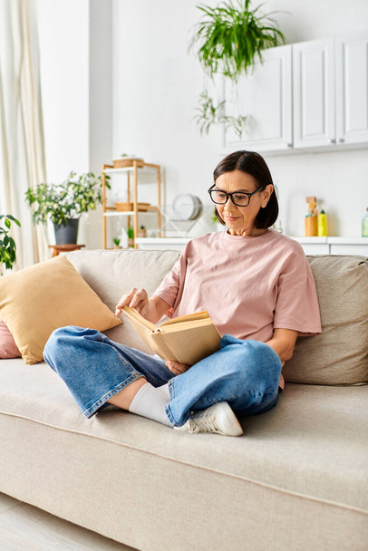 Dojrzała kobieta w przytulnym homewear siedzi na kanapie, w pełni zanurzona w czytaniu książki. - Zdjęcie, obraz