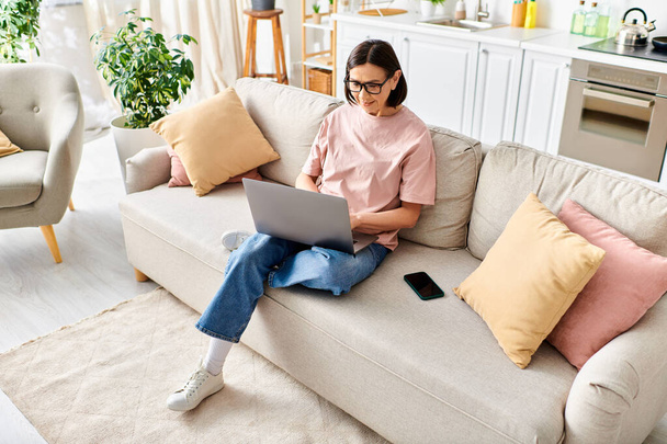 Взрослая женщина в уютной одежде сидит на диване и полностью сосредоточена на ноутбуке.. - Фото, изображение