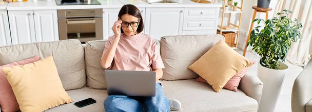 Uma mulher madura em roupa de casa aconchegante senta-se em um sofá, absorvida em usar um laptop. - Foto, Imagem