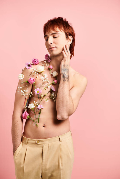 A shirtless young man strikes a pose with colorful flowers adorning his chest in a studio setting with a pink background - 写真・画像
