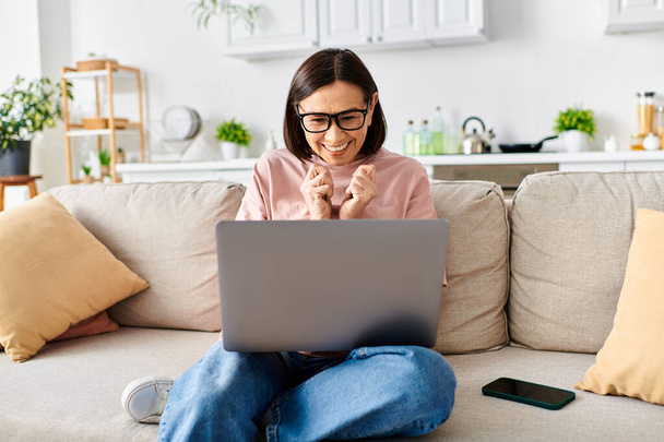 Dospělá žena v útulném oblečení sedí pohodlně na gauči, pohlcena svým laptopem. - Fotografie, Obrázek