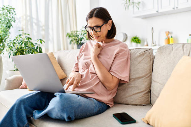 Reife Frau in gemütlicher Kleidung sitzt auf einer Couch, konzentriert auf einem Laptop-Bildschirm. - Foto, Bild