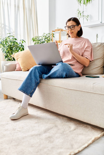 Een volwassen vrouw in gezellige huiskledij zittend op een bank, met behulp van een laptop. - Foto, afbeelding