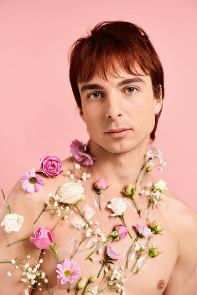 Młody człowiek bez koszuli pozuje z różnymi żywymi kwiatami zdobiącymi jego klatkę piersiową, ustawionymi na solidnym różowym tle. - Zdjęcie, obraz