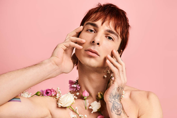 Ένας νεαρός άνδρας με τατουάζ στο στήθος ποζάρει με λουλούδια σε ένα στούντιο με ροζ φόντο.. - Φωτογραφία, εικόνα