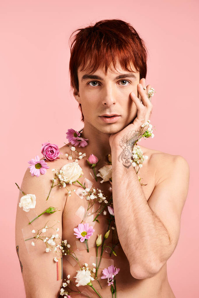 Młody mężczyzna przybiera pozę z kwiatami na klatce piersiowej na różowym tle w studio. - Zdjęcie, obraz