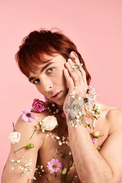 Νεαρός άνδρας με λουλούδια στο στήθος, κρατώντας τα χέρια στο πρόσωπο, στέκεται στο φόντο του στούντιο σε μια στάση αντανάκλασης. - Φωτογραφία, εικόνα
