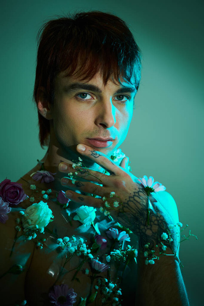 Egy fiatalember magabiztosan mutatja be a mellkasát színes tetoválásokkal és élénk virágokkal díszítve egy stúdióban, kék háttérrel.. - Fotó, kép