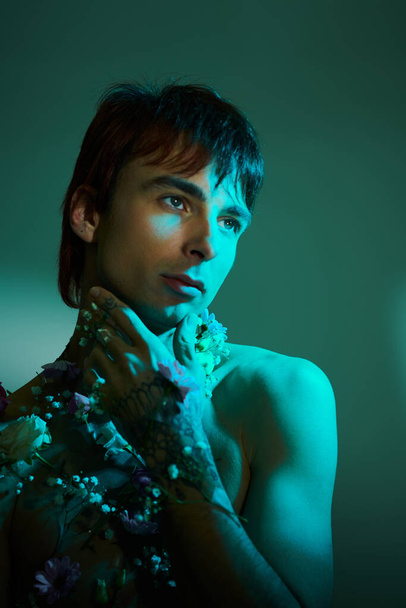 Un giovane uomo elegante in posa con i fiori intorno al collo in un ambiente studio con uno sfondo blu. - Foto, immagini