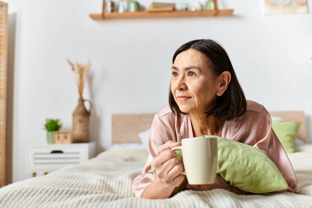 Een volwassen vrouw loungen op een bed met een kopje koffie in gezellige huiskledij. - Foto, afbeelding