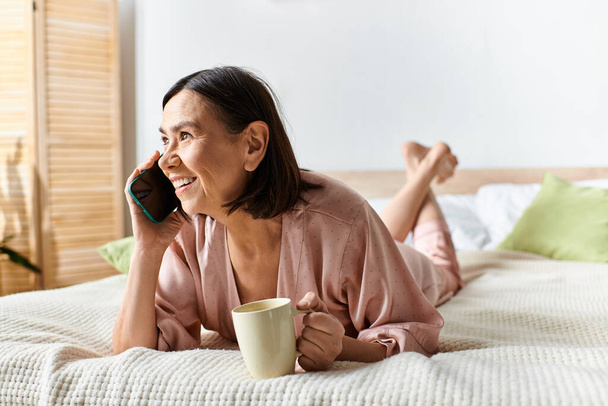 Una mujer en ropa de casa acogedora acostada en una cama, absorbida por una llamada telefónica. - Foto, Imagen