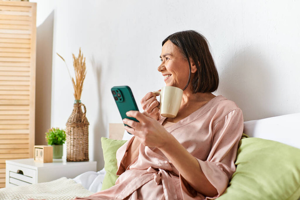 Femme en vêtements confortables jouit du café tout en vérifiant le téléphone sur le lit. - Photo, image