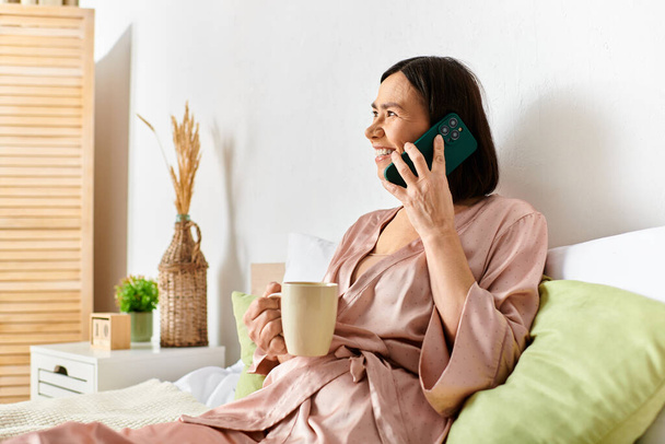 Ώριμη γυναίκα σε ζεστό homewear κάθεται χαλαρά στο κρεβάτι, βαθιά σε συνομιλία στο κινητό τηλέφωνο. - Φωτογραφία, εικόνα