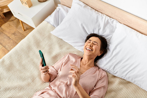 A mature woman in cozy homewear lays on a bed, holding a toothbrush. - Photo, Image