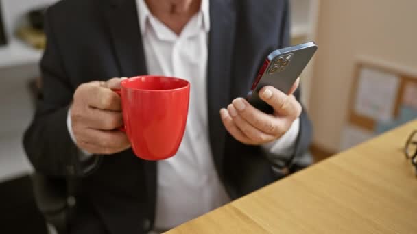 Starší šéf si vezme kávu, zabraný do kancelářské práce na svém smartphonu, obklopující portrét úspěchu - Záběry, video