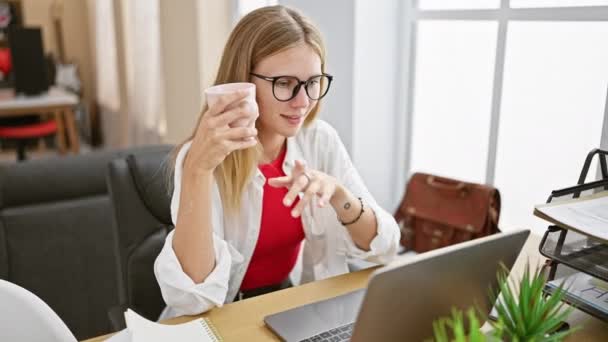 Mujer rubia en gafas sosteniendo una taza en un escritorio de la oficina con un ordenador portátil, portátil y una bolsa de cuero. - Imágenes, Vídeo