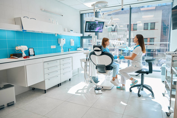 Стоматолог общается с молодым пациентом в стоматологическом кабинете, врач держит девушек за руки - Фото, изображение