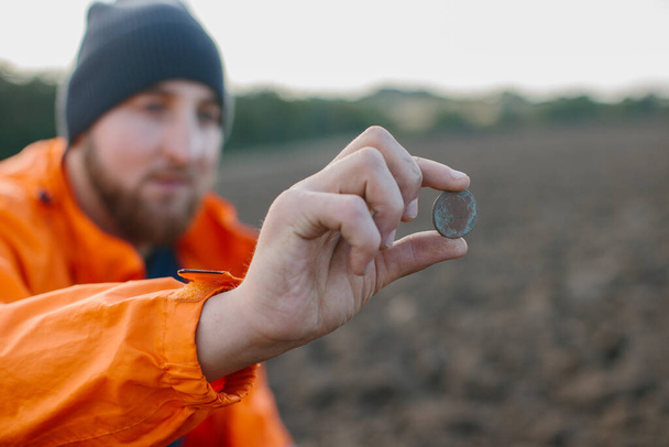Un hombre tiene una moneda antigua en sus manos, encontrada en un campo con un detector de metales. - Foto, imagen