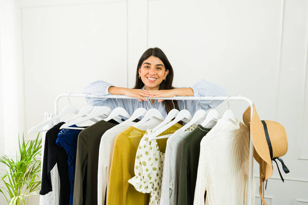 Fröhliche weibliche Mode-Enthusiastin posiert mit einer Vielzahl stylischer Outfits auf einem Kleiderständer - Foto, Bild