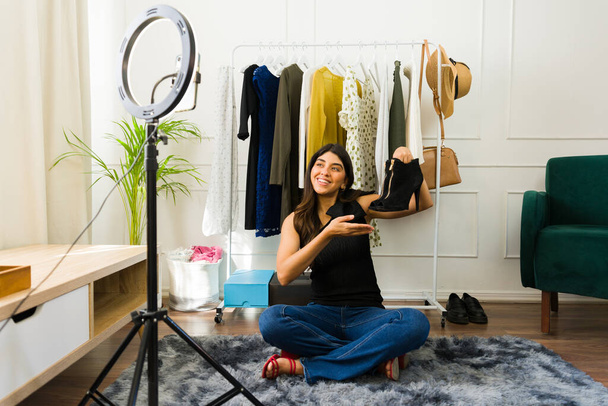 Молодая женщина vlogging о моде, сидя возле вешалки одежды и улыбаясь на камеру - Фото, изображение