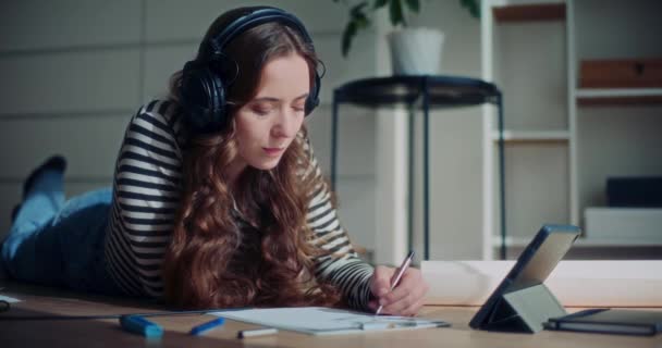 Pretty młoda brunetka leżąca na podłodze podczas rysowania i słuchania muzyki przez słuchawki w salonie w domu - Materiał filmowy, wideo