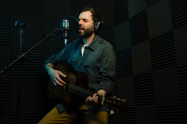 Férfi művész szenvedélyesen gitározik és énekel a mikrofonba egy hangszigetelt stúdióban - Fotó, kép