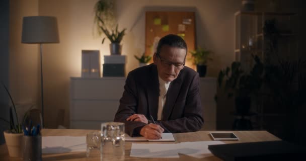 Hombre de negocios sénior pensando mientras escribe en el diario en la mesa tarde en la noche en la oficina - Imágenes, Vídeo