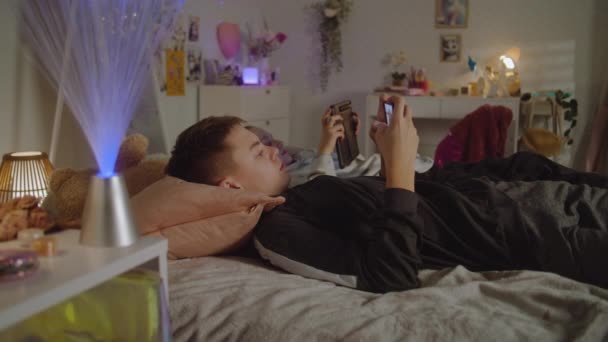 Fiatal lány fekszik az ágyon a szobában, és néz videó tartalmat a tabletta. A fiú telefonnal a kezében videojátékozik. A tinédzserek szabadidőt töltenek. Családi kapcsolat. Otthon hangulatos elegáns belső. - Felvétel, videó