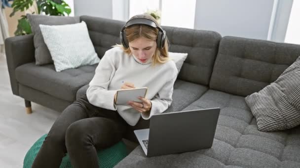 A blonde woman works from home, seated on a pouf with headphones, a laptop, writing in a notebook, in a modern living room. - Footage, Video