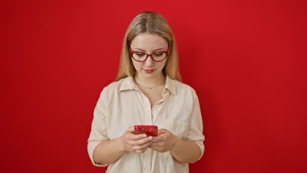 Mujer rubia joven vivaz clava la alegría de un texto amistoso sobre la pared roja de pie con confianza con una sonrisa dentada y un teléfono inteligente - Imágenes, Vídeo