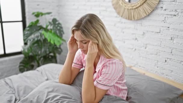 Joven mujer caucásica que aparece estresada y fatigada sentada en su dormitorio con una pared de ladrillo blanco en el fondo. - Metraje, vídeo
