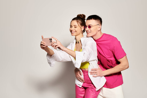 Młoda stylowa para uśmiecha się podczas robienia selfie z telefonem komórkowym w studiu na szarym tle. - Zdjęcie, obraz