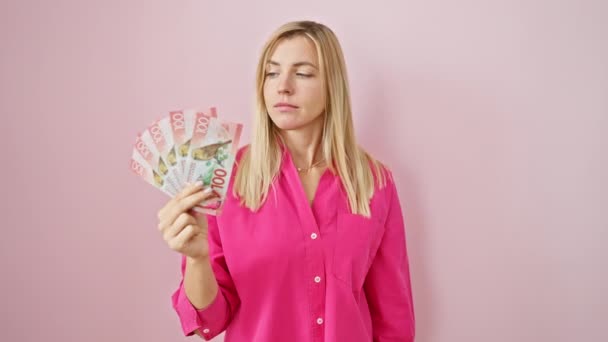 Молодая блондинка, держащая в руках доллары Новой Зеландии, указывая пальцем на камеру и на вас, уверенный в себе жест, выглядящий серьезным на изолированном розовом фоне - Кадры, видео