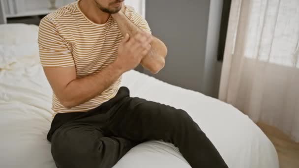 Homme hispanique appliquant une lotion au coude dans un cadre confortable de chambre à coucher, transmettant une routine de soins personnels. - Séquence, vidéo