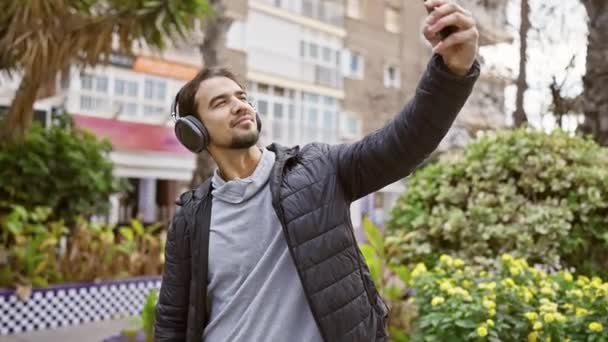 Een jonge Spaanse man met een koptelefoon neemt een selfie in een levendige stadstuin. - Video
