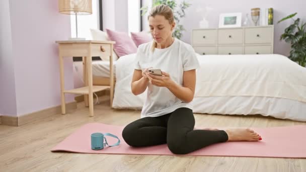 Junge Frau sitzt in ihrem Schlafzimmer auf einer Yogamatte und benutzt ein Smartphone mit einem Lautsprecher neben sich - Filmmaterial, Video