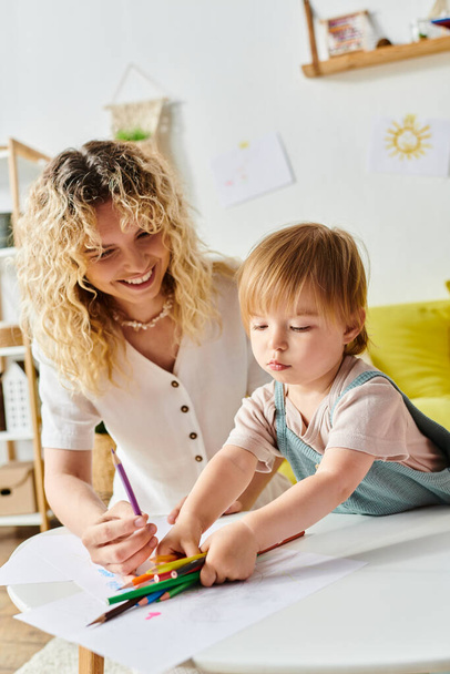 Curly-haired mother gently guides her toddler daughters hand while drawing with colorful crayons at home. - Photo, Image
