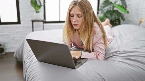 Szőke nő fekszik az ágyon laptoppal egy modern hálószobában természetes fény és növények - Felvétel, videó