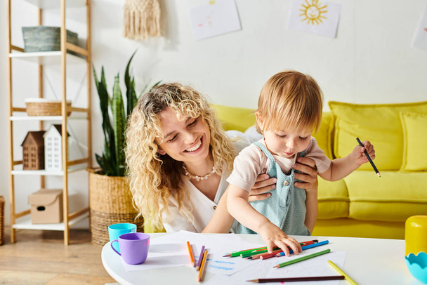 Kıvırcık saçlı bir anne ve küçük kızı, yaratıcı bir ifadeyle meşgul olan pastel boyalarla çevrili bir masada oturuyorlar.. - Fotoğraf, Görsel