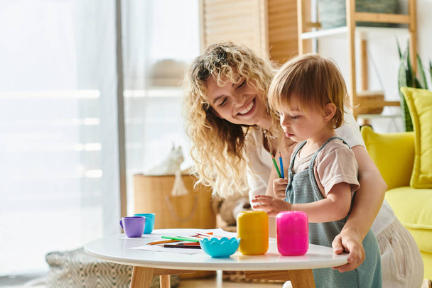 Μια σγουρομάλλα μητέρα και το παιδί της κόρη παιχνιδιάρικα διερευνήσει και να μάθουν, ενώ χρησιμοποιώντας κύπελλα σε μια δραστηριότητα εμπνευσμένη από Montessori στο σπίτι. - Φωτογραφία, εικόνα