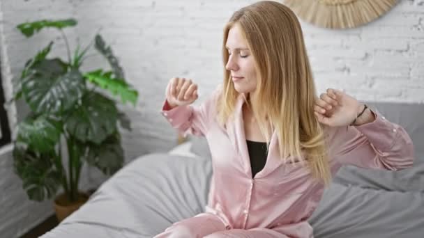 Vonzó fiatal nő rózsaszín pizsamában nyújtózkodik, miután felébredt a stílusos hálószobájában. - Felvétel, videó
