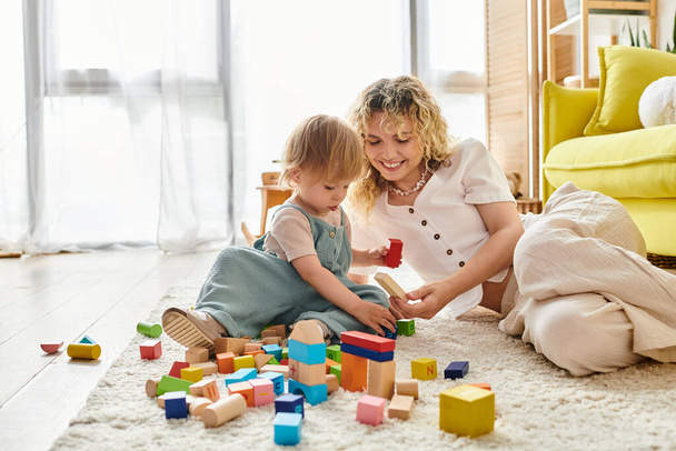 A curly mother and toddler daughter enjoy a playful moment on the floor, building structures with colorful blocks. - Photo, Image