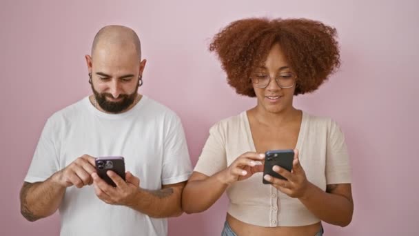 Krásný pár usmívá, stojí spolu nad růžové izolované pozadí, těší sms na smartphonech, vyzařuje důvěru a lásku - Záběry, video