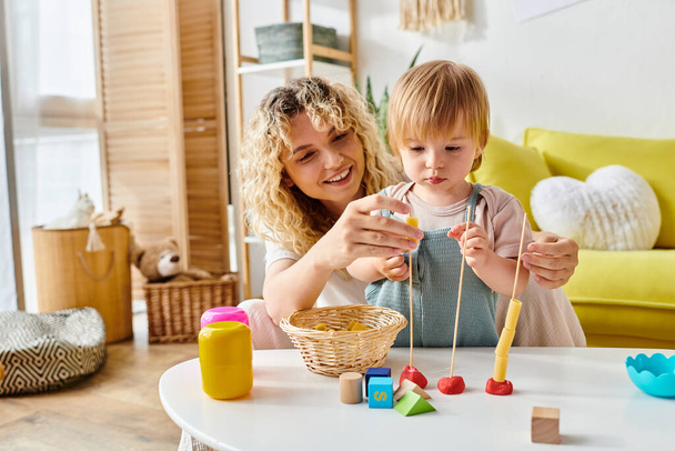 Μια σγουρή μητέρα και η κόρη της συμμετέχουν σε παιχνιδιάρικη μάθηση με ξύλινα παιχνίδια στο σπίτι, απολαμβάνοντας τη μέθοδο της εκπαίδευσης Montessori. - Φωτογραφία, εικόνα