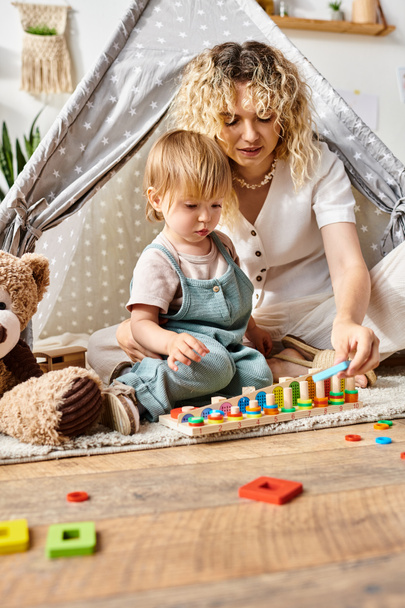 Una madre de pelo rizado y su hija pequeña juegan alegremente cerca del oso de peluche, participando en la educación imaginativa inspirada en Montessori en el hogar.. - Foto, imagen