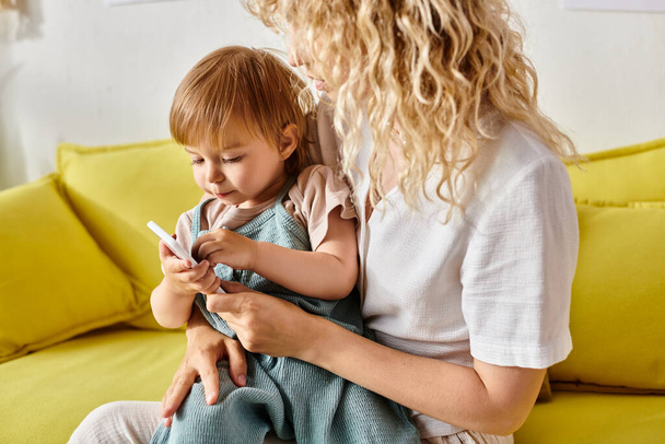 Mãe de cabelos encaracolados segurando a filha da criança que é hipnotizada por uma tela de telefone celular em casa. - Foto, Imagem