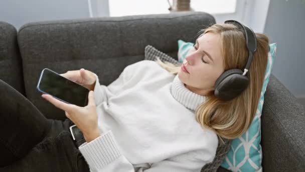 Mladá žena se sluchátky relaxuje na gauči pomocí chytrého telefonu, ztělesňuje volný čas v útulném obývacím pokoji. - Záběry, video