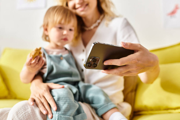 Μια σγουρή μητέρα κρατά ένα κινητό δίπλα στην κόρη της στο σπίτι, που ασχολείται με μια συγκινητική στιγμή σύνδεσης.. - Φωτογραφία, εικόνα