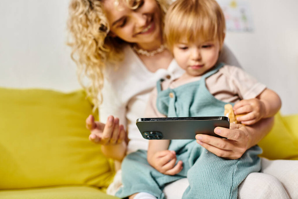 A curly mother attentively holds her toddler daughter while looking at a tablet in a cozy home setting. - Photo, Image